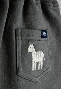 Grå stoflomme med en broderet hvid zebra og en lille marineblå mærkat med mærkenavnet "JoJo" fastgjort øverst.