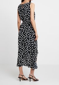 Robe noire à pois blancs avec un design sans manches, taille cintrée, dos avec ouverture en forme de goutte et ourlet fluide. Associée à des sandales noires à lanières.
