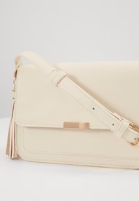 Krémová kožená kabelka přes rameno s klopou na zavírání, zlatými kovovými detaily, nastavitelným popruhem a ozdobnou střapcem na boku.