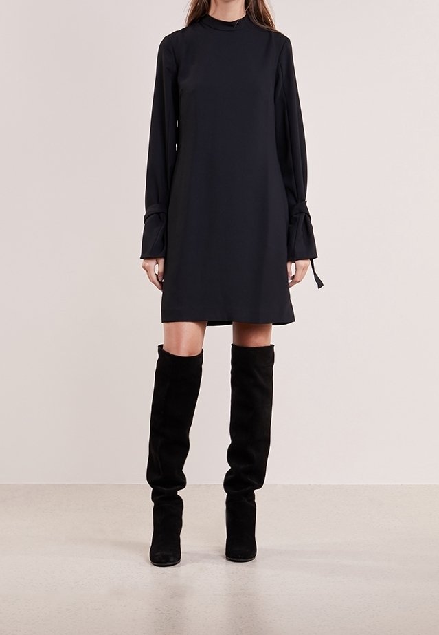 Robe noire à manches longues avec un col montant et des poignets noués, associée à des bottes en daim noires jusqu'aux genoux, créant une silhouette élégante.