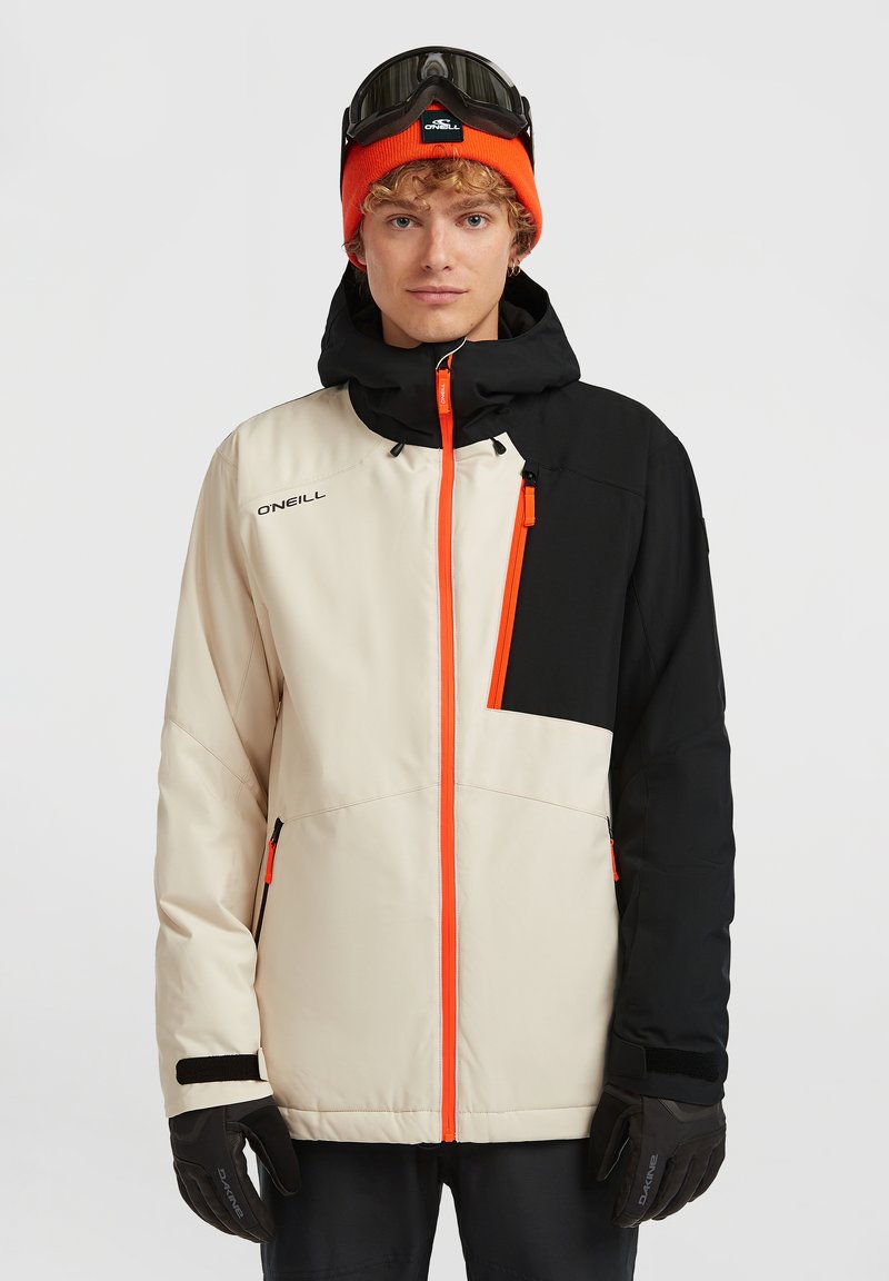 Jeune homme portant une veste de ski noire et beige avec des fermetures éclair orange, des gants noirs, un bonnet orange et des lunettes de ski noires sur la tête.