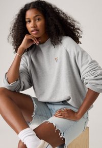 Felpa grigia con un logo triangolare colorato, vestibilità comoda, polsini a costine, indossata con pantaloni corti in denim chiaro strappati.