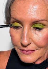 Close-up van een model die felgroene oogschaduw aanbrengt met een kwast. De huid is glad met een natuurlijke afwerking; de lippen zijn getint in licht mauve.
