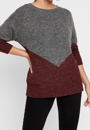 Femme portant un pull à manches longues bicolore avec une partie supérieure grise et une partie inférieure bordeaux, associé à un pantalon noir.