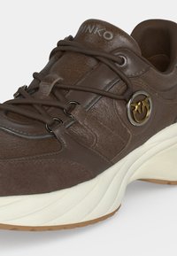 Baskets en cuir marron avec semelle blanche épaisse, emblème rond avec logo représentant deux oiseaux et lacets croisés sur fond clair.