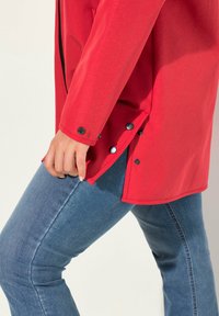 Giacca di tessuto intrecciato rosso con taglio dritto, bottoni laterali all'orlo e una mano parzialmente nascosta vicino a un pantalone in denim blu.