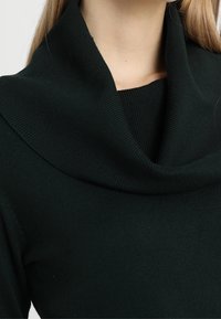 Pull en maille vert foncé avec un large col châle plié et une texture côtelée, présentant une silhouette ajustée.