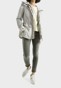 Parka ligera de color gris claro, con capucha ajustable, cintura elástica y dos bolsillos laterales, combinada con pantalones verde olivo y zapatillas blancas.