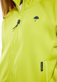 Ljusgul jacka med strukturerad tyg, utrustad med dragkedja och en liten blå paraplylogotyp. Inkluderar en ficka med dragkedja med varumärke.