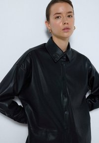 Černá kožená košile-jacket s knoflíkovým zapínáním, špicatým límcem a volným střihem, s jemným leskem a měkkou texturou.