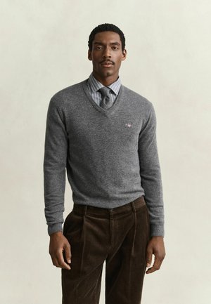 Uomo che indossa un maglione grigio a scollo a V sopra una camicia a righe e cravatta, abbinato a pantaloni di velluto a coste marroni, in piedi davanti a uno sfondo semplice.