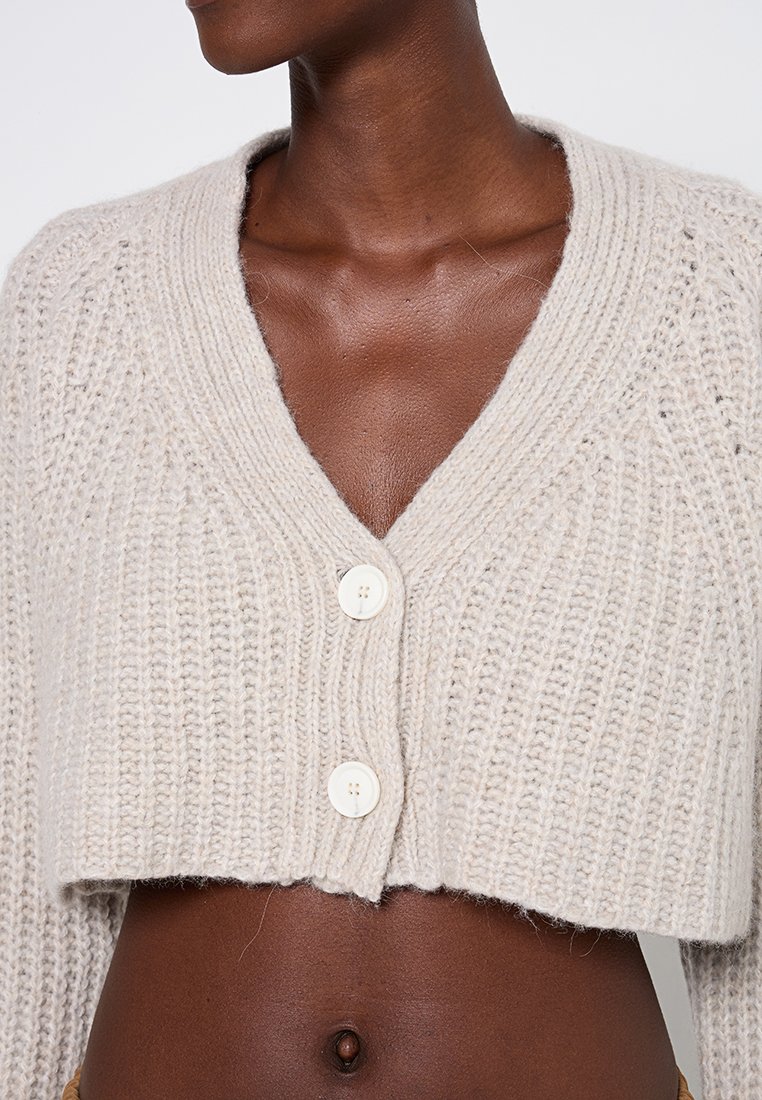 Häkeljacke in hellbeige mit V-Ausschnitt, drei Knöpfen und figurbetontem Schnitt. Strukturstrick mit Rippdetails.