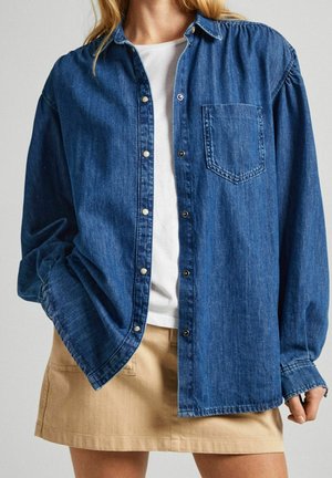 Camicia di jeans denim di colore blu scuro con maniche lunghe, una tasca sul petto e bottoni a pressione argentati; abbinata a una gonna beige.