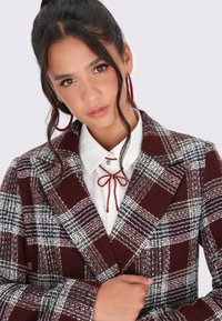 Karierter Blazer in Burgunderrot, Weiß und Grün. Strukturierten Stoff mit taillierter Passform und Revers. Begleitet von einer weißen Bluse und roten Akzenten.