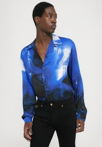 Camicia a maniche lunghe con bottoni di un blu vivace con motivi astratti, realizzata in tessuto morbido, abbinata a jeans neri aderenti con accenti dorati.