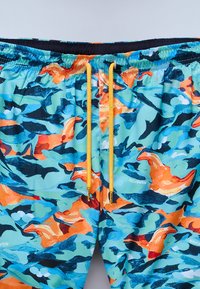 Bañador con un vibrante patrón de camuflaje multicolor en azul, naranja y negro. Cuenta con una cinturilla elástica y cordones amarillos.