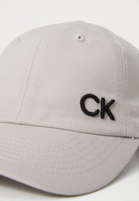 Világosszürke baseball sapka pamut keverékből, amelyen elöl fekete hímzett "CK" logó és textúrázott varrásminták találhatóak.