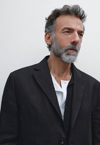 Schwarzer, taillierter Blazer mit subtiler Textur, getragen über einem hellen weißen Button-Down-Hemd mit einem einzigartigen Kragen und Designelementen.