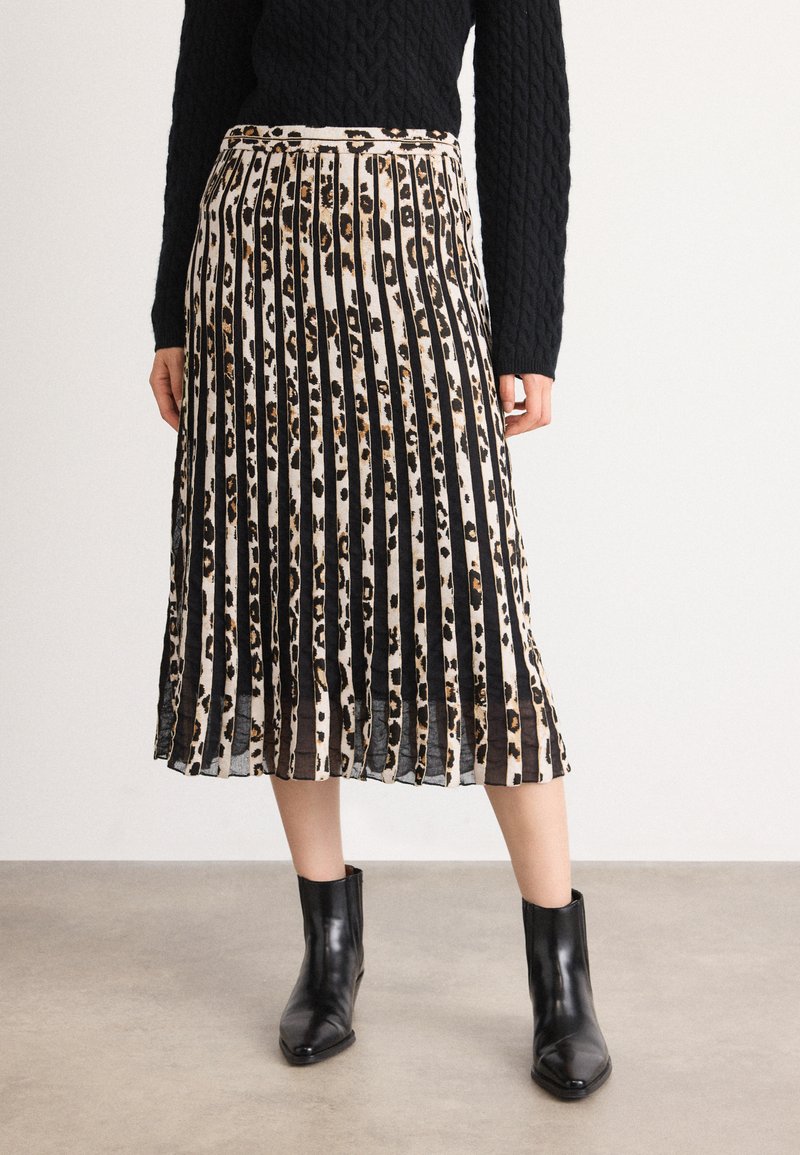 Vrouw in een geplooide midi rok met zwart-beige luipaardprint, een zwart kabelgebreid trui en zwarte enkellaarsjes, staande op een grijze vloer.