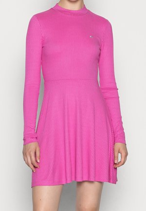 Femme portant une robe rose vif à manches longues côtelées avec un col montant, debout devant un fond clair uni.