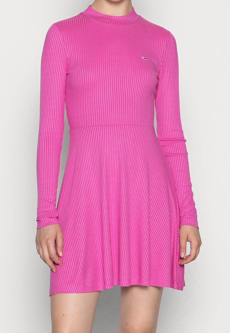 Femme portant une robe rose vif à manches longues côtelées avec un col montant, debout devant un fond clair uni.