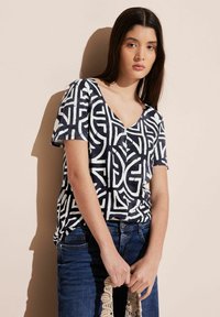 Navy und weiß gemusterte T-Shirt mit V-Ausschnitt und kurzen Ärmeln, das geometrische Designs aufweist. Kombiniert mit blauen Jeans und einer Makramee-Tasche.