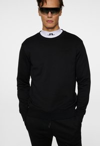 Felpa nera con colletto rotondo dalla finitura testurizzata, polsini e orlo a coste, con un discreto dettaglio del logo sul petto. Occhiali da sole neri indossati.