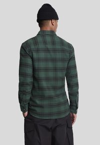 Camicia di flanella a quadri verdi con accenti neri, maniche lunghe, polsini con bottoni e orlo curvo. Indossata con pantaloni cargo neri, vista dal retro.