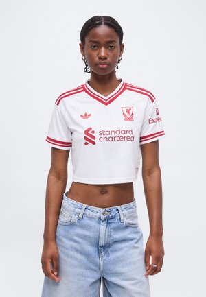 Femme aux cheveux tressés portant un maillot de football blanc court du Liverpool FC avec des logos rouges et un jean bleu clair, debout devant un fond uni.