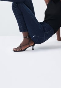 Sandalias de tacón negro con tiras y diseño de punta cuadrada, combinadas con unos jeans ajustados de color azul oscuro, mostrando una camiseta de textura acanalada.