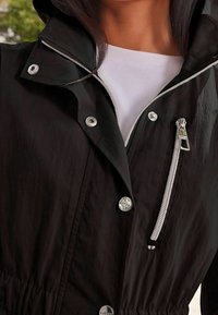 Chaqueta ligera negra con capucha, cuenta con cremallera plateada y botones a presión, cintura fruncida y una capa interior blanca a contraste.