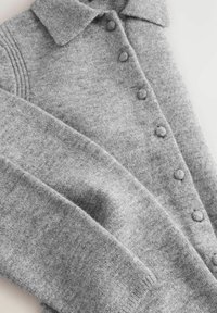 Cardigan tricoté gris doux avec boutons recouverts de tissu, manches longues et col, posé à plat sur une surface blanche.