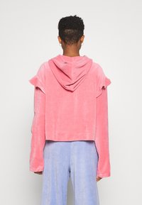 Sudadera rosa de terciopelo cortada con un diseño acampanado en las mangas y capucha. Textura suave con un ajuste relajado. Combinada con pantalones de textura azul claro.