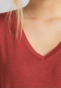 Gros plan sur une personne portant un t-shirt rouge en V, révélant le décolleté et une partie de ses cheveux blonds sur l'épaule.