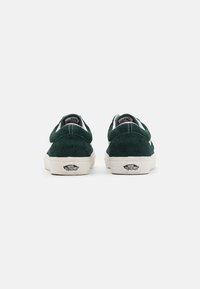 Grüne Wildleder-Sneaker im Low-Top-Design mit weißen Akzenten und einer Gummisohle, die ein Logo an der Ferse trägt. Das Design ist schlicht und funktional.