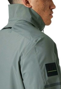 Chaqueta impermeable gris con cuello alto, tejido texturizado y parche de logo negro en el brazo izquierdo. Presenta detalles reflectantes en los hombros.