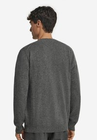 Mężczyzna odwrócony plecami, ubrany w ciemnoszary sweter dzianinowy z długimi rękawami i prążkowanymi mankietami, na jednolitym białym tle.