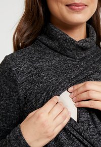 Grauer Rollkragenpulli mit strukturiertem Strickmuster, ausgestattet mit einer unauffälligen Öffnung an der Vorderseite, die eine hellere innere Schicht enthüllt.