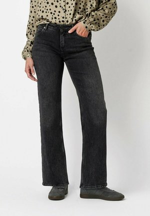 Person, die schwarze Jeans mit weitem Bein, graue Turnschuhe und ein beiges Langarmshirt mit schwarzen Punkten trägt, steht mit einer Hand in der Tasche.