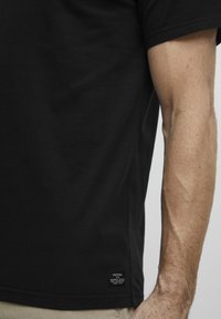 Juodas medvilnės marškinėlis su etikete kairiajame apatiniame kampe, trumpomis rankovėmis ir apvaliu kaklu. Lygus audinys su minimaliais dizaino akcentais.