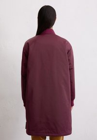 Lange, maroon pufferjack met geribbelde kraag en manchetten, gladde textuur en zijzakken. Bereikt de lengte tot halverwege de dijen.