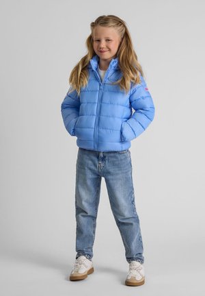 Ung flicka står med händerna i fickorna, klädd i en ljusblå puffjacka, blå jeans och vita sneakers framför en neutral bakgrund.