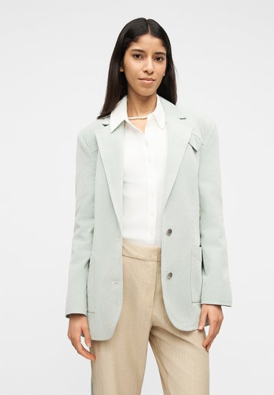 Γυναίκα με μακριά σκούρα μαλλιά, που φοράει ανοιχτό γκρι σακάκι, λευκό πουκάμισο με κουμπιά, μπεζ παντελόνι και κολιέ με πέρλες, στέκεται μπροστά σε απλό φόντο.