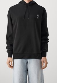 Černá mikina s kapucí z měkké látky, s šňůrkou na kapuci, žebrovanými manžetami a malou grafikou Tokia na hrudi. Kombinováno se světle modrými džíny.