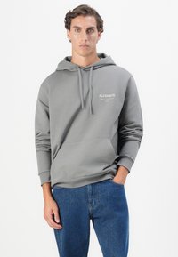 Grå hettegenser laget av mykt stoff med kengurulommer, justerbare snorene og en hvit trykt logo på brystet. Brukt med blå jeans.