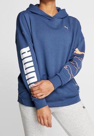 Persoon draagt een blauw Puma-hoodie met een wit logo op de linkerborst, grote witte "PUMA" tekst op de rechtermouw en oranje Puma-kattooutline op de linkermouw.