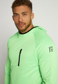 Neon grøn hættejakke med hætte og lynlåslomme på brystet. Har sorte accenter omkring lommen og ærmet. Glat, letvægtsstof.