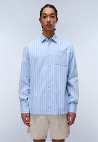 Camisa a rayas azul claro hecha de tela suave, con un bolsillo frontal, botones y mangas largas. Llevada con pantalones cortos beige.
