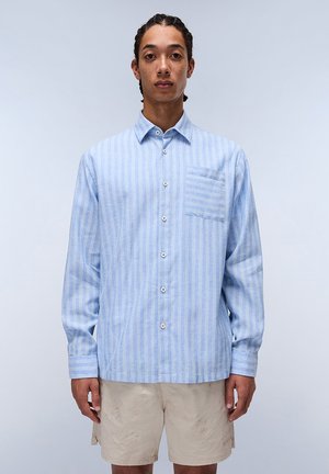 Camisa a rayas azul claro hecha de tela suave, con un bolsillo frontal, botones y mangas largas. Llevada con pantalones cortos beige.