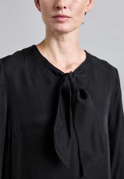 Black blouse featuring a round neckline, tie detail at the front, and smooth texture. The fabric appears lightweight with a soft drape.